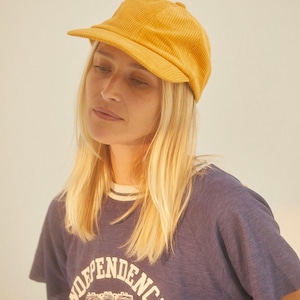 Könnte beinhalten: Eine Frau trägt ein blaues T-Shirt mit einem weißen "Independence"-Aufdruck und eine gelbe Baseballkappe aus Cord.
