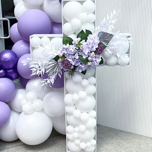 May include: A large white cross-shaped decoration filled with white balloons. The cross is adorned with a bouquet of purple and white flowers and greenery. A cluster of purple and white balloons is on the left.