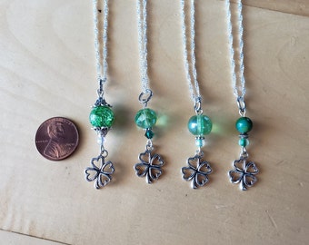 May include: Four silver necklaces with green beads and clover pendants. The beads vary in shape and texture, with one having a glittery appearance. A penny is included for size reference. The necklaces are displayed on a light brown surface.