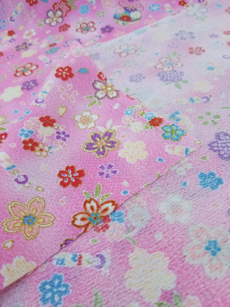 May include: Pink fabric with a floral pattern. The fabric features a variety of pink, white, blue, and yellow flowers. The fabric is folded in half, showing the pattern on both sides.