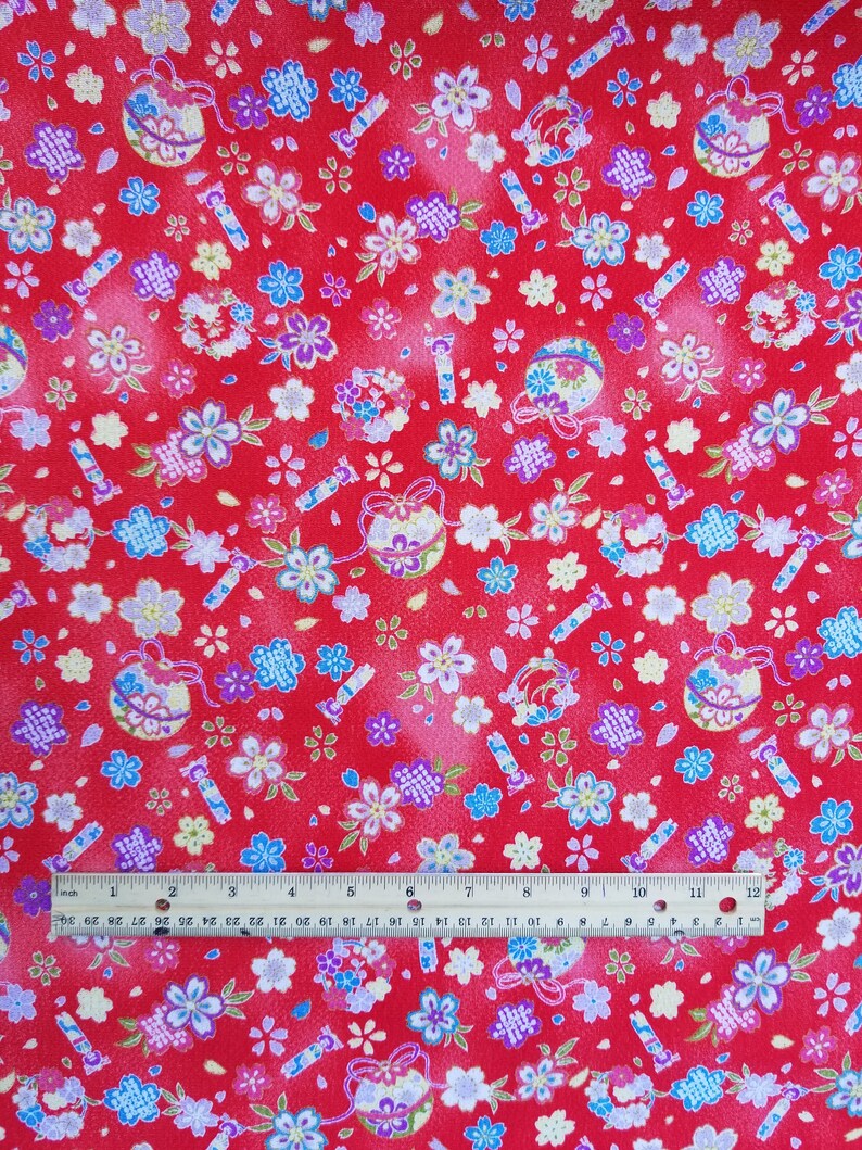 May include: Red fabric with a repeating pattern of small white, blue, and pink flowers, gold and silver ornaments, and red and gold sticks. The fabric is measured with a ruler in inches.