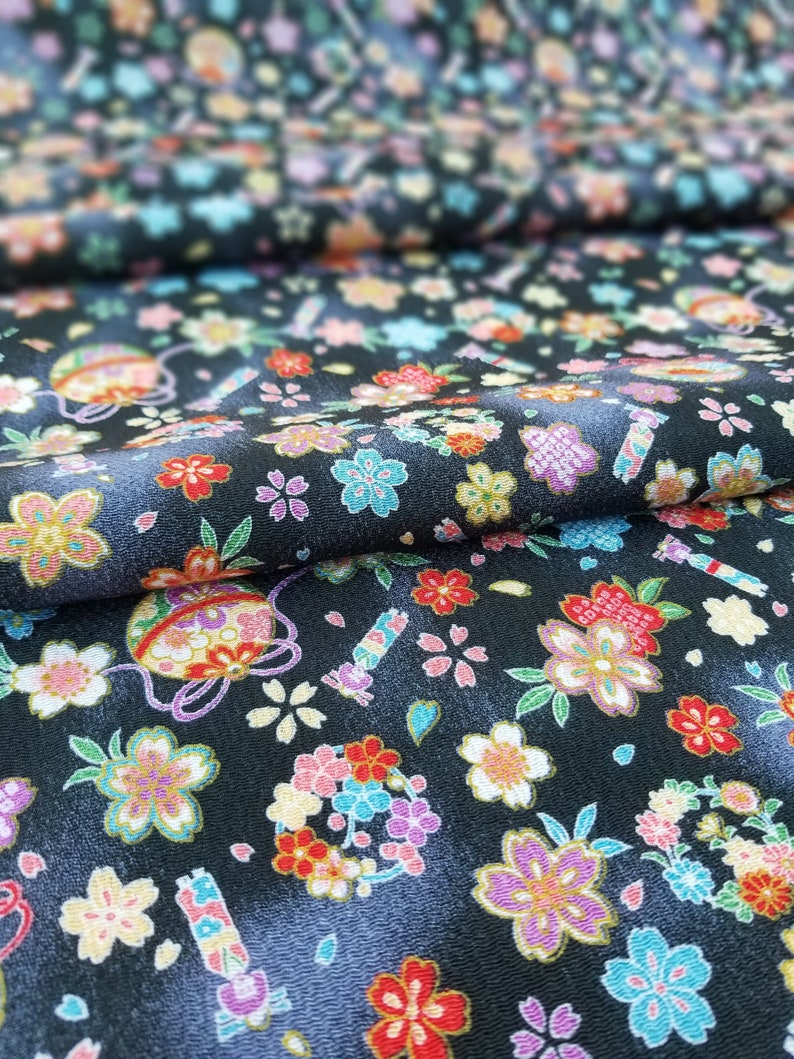 May include: Black fabric with a repeating pattern of colorful flowers and small ornaments. The fabric has a subtle sheen and a textured surface.