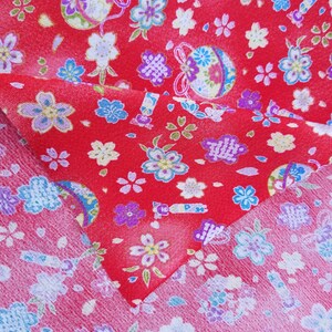 May include: Red fabric with a repeating pattern of white and blue flowers and a small, colorful, round ornament. The fabric is folded in half, showing the pattern on both sides.