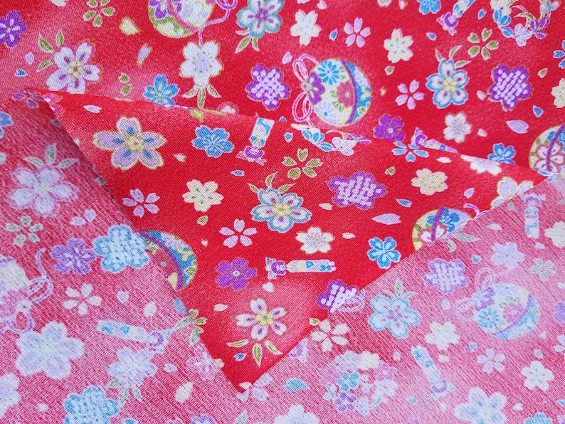 May include: Red fabric with a repeating pattern of white and blue flowers and a small, colorful, round ornament. The fabric is folded in half, showing the pattern on both sides.