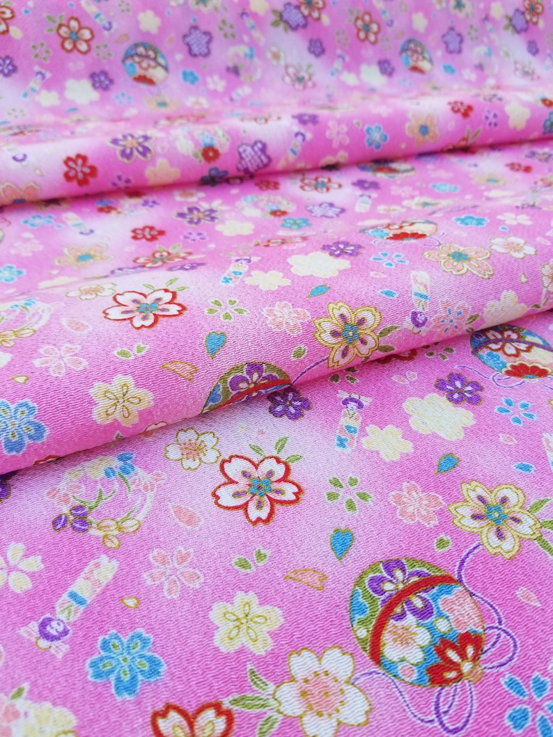 May include: Pink fabric with a repeating pattern of small flowers and a larger, colorful, round design. The fabric has a subtle ombre effect, fading from a darker pink to a lighter pink.