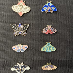 May include: Collection of enamel moth pins in various colors, including pink, blue, black, and green. Each pin features intricate details and celestial designs, perfect for adding a touch of whimsy to jackets, bags, or hats.