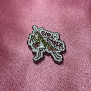 May include: A white and green enamel pin featuring a praying mantis with the words "Girl Dinner". The pin has pink and white accents and is set against a pink background.