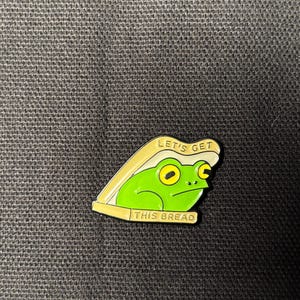May include: A green frog pin with yellow eyes and a white and tan bread crust. The pin has the text "LET'S GET THIS BREAD" in white letters. The pin is on a black textured surface.