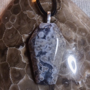 Könnte beinhalten: Ein Edelsteinanhänger mit einem einzigartigen Muster, an einer schwarzen Kordel befestigt. Der Anhänger zeigt eine Mischung aus Grau-, Weiß- und Blautönen mit polierter Oberfläche. Der Hintergrund ist ein strukturierter, brauner Stein, der die natürliche Schönheit des Anhängers hervorhebt. Ein stilvolles Accessoire.
