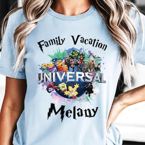 May include: Light blue t-shirt featuring a colorful graphic with the text "Family Vacation UNIVERSAL Melany." The graphic includes various cartoon characters. The shirt is made of a soft material.
