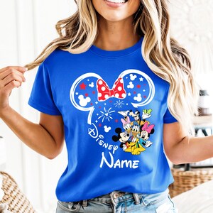 Puede incluir: Camiseta azul real con un dise&ntilde;o de oreja de Minnie Mouse blanca, un lazo rojo de lunares y fuegos artificiales. La camiseta incluye un gr&aacute;fico de personajes de Disney y la palabra "Disney" con espacio para un nombre.