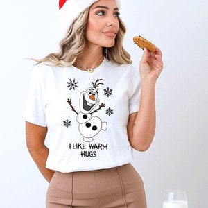 May include: White short-sleeve t-shirt featuring a cartoon snowman character and snowflakes. The text "I LIKE WARM HUGS" is printed below the snowman. The person in the image is wearing a Santa hat.