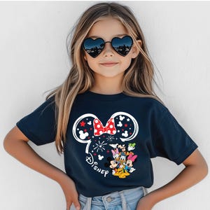 Puede incluir: Camiseta azul marino con un dise&ntilde;o de Minnie Mouse. El dise&ntilde;o incluye un contorno blanco de la cabeza de Minnie con un lazo rojo, estrellas y la palabra "Disney". Varios personajes de Disney est&aacute;n debajo del lazo.