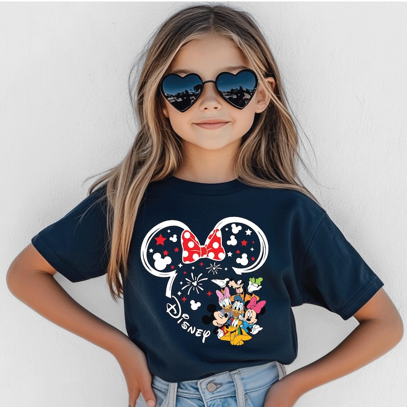 Puede incluir: Camiseta azul marino con un dise&ntilde;o de Minnie Mouse. El dise&ntilde;o incluye un contorno blanco de la cabeza de Minnie con un lazo rojo, estrellas y la palabra "Disney". Varios personajes de Disney est&aacute;n debajo del lazo.