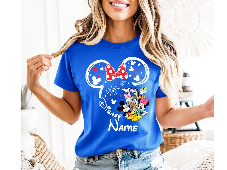 Puede incluir: Camiseta azul real con un dise&ntilde;o de oreja de Minnie Mouse blanca, un lazo rojo de lunares y fuegos artificiales. La camiseta incluye un gr&aacute;fico de personajes de Disney y la palabra "Disney" con espacio para un nombre.