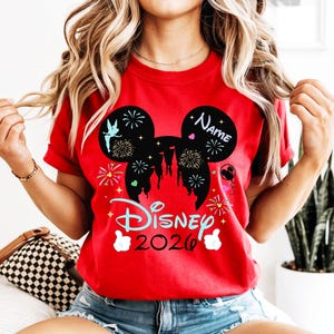 May include: A red t-shirt with a black Mickey Mouse silhouette design, fireworks, and a castle. The shirt displays "Disney 2026" and a space for a name. Two thumbs up are at the bottom.