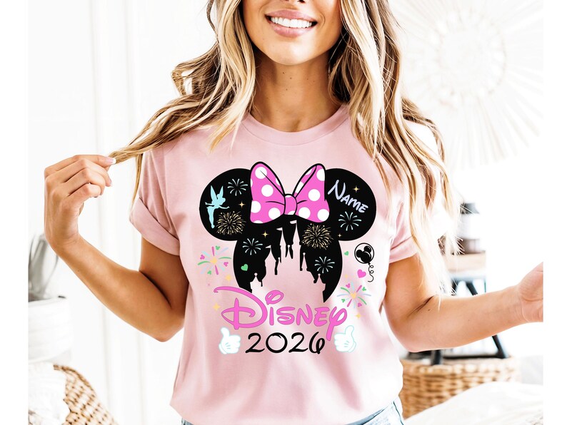 May include: A pale pink t-shirt with a black Minnie Mouse design, including a pink polka dot bow. The design features fireworks, a castle silhouette, and the word "Disney" with the year "2026". The word "Name" is also included.