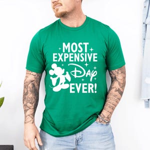 May include: A green crew neck t-shirt featuring the white text "MOST EXPENSIVE Day EVER!" and a white Mickey Mouse silhouette. The shirt is a vibrant green colour.