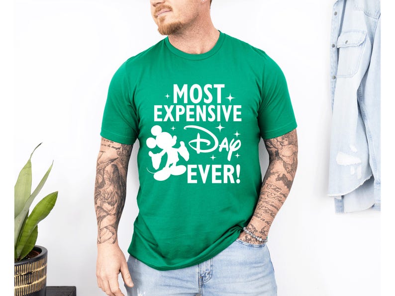 May include: A green crew neck t-shirt featuring the white text "MOST EXPENSIVE Day EVER!" and a white Mickey Mouse silhouette. The shirt is a vibrant green colour.