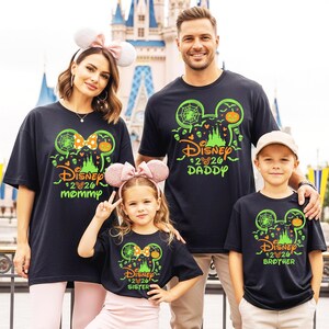 May include: Black short-sleeve t-shirts featuring a neon green and orange graphic of a Mickey Mouse silhouette, a castle, spiderwebs, and pumpkins. The text reads Disney 2026 with individual labels including Mommy, Daddy, Sister, and Brother.