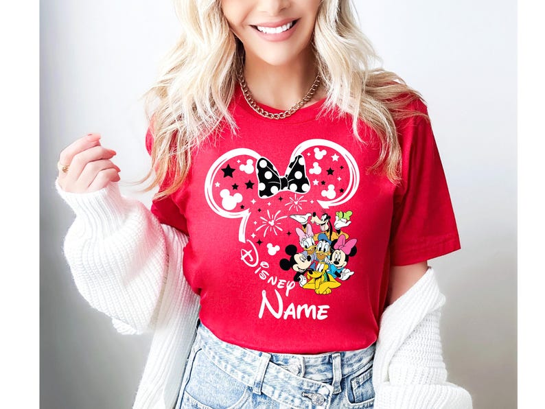 Puede incluir: Camiseta roja con un dise&ntilde;o tem&aacute;tico de Disney. El dise&ntilde;o incluye una silueta de Minnie Mouse con un lazo de lunares blancos y negros, estrellas y un grupo de personajes de Disney. Tambi&eacute;n es visible el texto "Disney NAME".