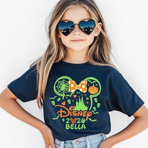 May include: A dark blue short-sleeve t-shirt featuring a neon green graphic of Mickey Mouse ears, a pumpkin, a spider web, and a castle. The design includes an orange polka-dot bow, the word Disney, the year 2026, and the name Bella in neon green.