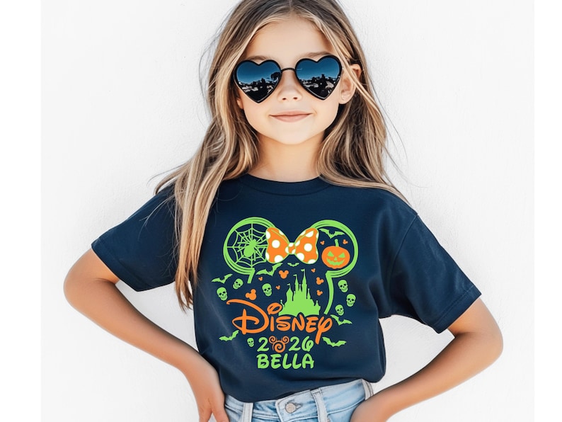 May include: A dark blue short-sleeve t-shirt featuring a neon green graphic of Mickey Mouse ears, a pumpkin, a spider web, and a castle. The design includes an orange polka-dot bow, the word Disney, the year 2026, and the name Bella in neon green.