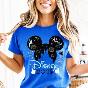 May include: A royal blue t-shirt with a black Mickey Mouse silhouette design, including fireworks, a castle, and a fairy. The shirt displays "Disney 2026" and a space for a name. White gloves are at the bottom of the design.