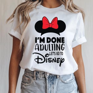 May include: White t-shirt with a black Minnie Mouse ears silhouette and a red bow. The text "I'M DONE ADULTING Let's Go To Disney" is printed in black. A casual wear item, suitable for a trip to a theme park.