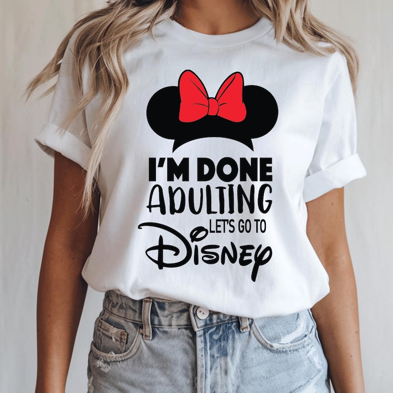 May include: White t-shirt with a black Minnie Mouse ears silhouette and a red bow. The text "I'M DONE ADULTING Let's Go To Disney" is printed in black. A casual wear item, suitable for a trip to a theme park.