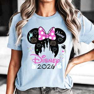 May include: Pale blue t-shirt with a Minnie Mouse design, including a pink polka dot bow, fireworks, and a castle silhouette. The text "Disney 2026" is in pink, with space for a name. A cheerful and celebratory design.