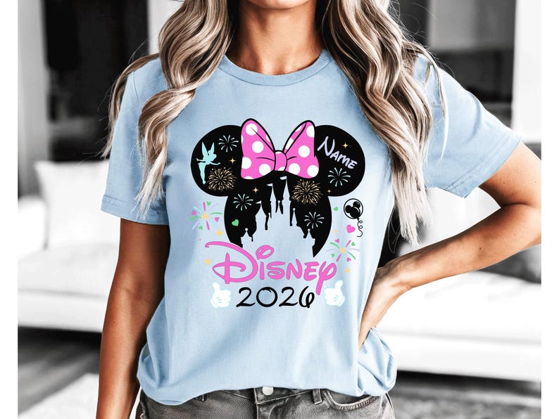 May include: Pale blue t-shirt with a Minnie Mouse design, including a pink polka dot bow, fireworks, and a castle silhouette. The text "Disney 2026" is in pink, with space for a name. A cheerful and celebratory design.