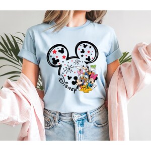 Puede incluir: Camiseta azul claro con un dise&ntilde;o de cabeza de Mickey Mouse negra con estrellas rojas y fuegos artificiales. La palabra "Disney" est&aacute; impresa debajo. La camiseta tambi&eacute;n presenta un gr&aacute;fico colorido de personajes de Disney.