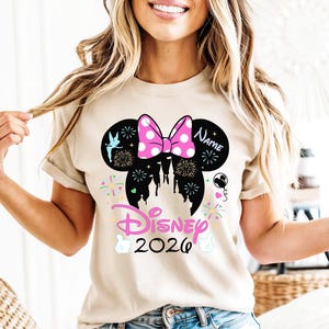 May include: A beige t-shirt with a black Mickey Mouse ears silhouette, a pink polka-dot bow, fireworks, and a castle design. The word "Disney" and the year "2026" are also featured.