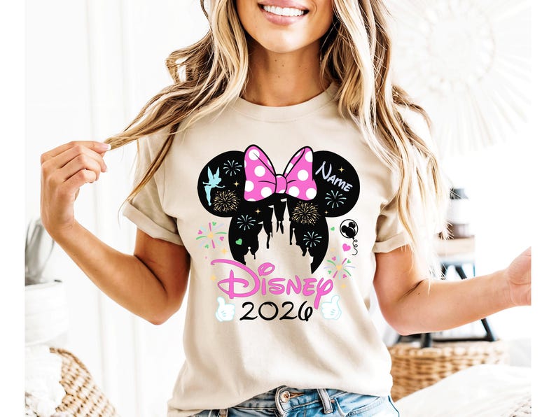 May include: A beige t-shirt with a black Mickey Mouse ears silhouette, a pink polka-dot bow, fireworks, and a castle design. The word "Disney" and the year "2026" are also featured.
