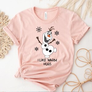 Peut inclure: T-shirt rose clair avec un dessin de bonhomme de neige de dessin anim&eacute; et le texte "I LIKE WARM HUGS". Le bonhomme de neige a un nez en carotte, des bras en b&acirc;ton et des flocons de neige.