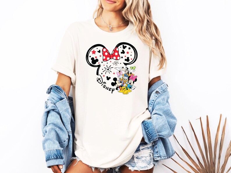 Puede incluir: Camiseta color crema con un contorno negro de las orejas de Mickey Mouse, un lazo rojo y estrellas. La camiseta tiene un gr&aacute;fico de Disney con Mickey, Minnie, Donald, Daisy y Goofy. La modelo lleva una chaqueta vaquera.