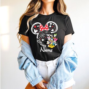 Puede incluir: Camiseta negra con un dise&ntilde;o de Minnie Mouse, un lazo rojo y el texto "Disney Name". El dise&ntilde;o incluye fuegos artificiales y personajes de Disney. La camiseta tiene cuello redondo.
