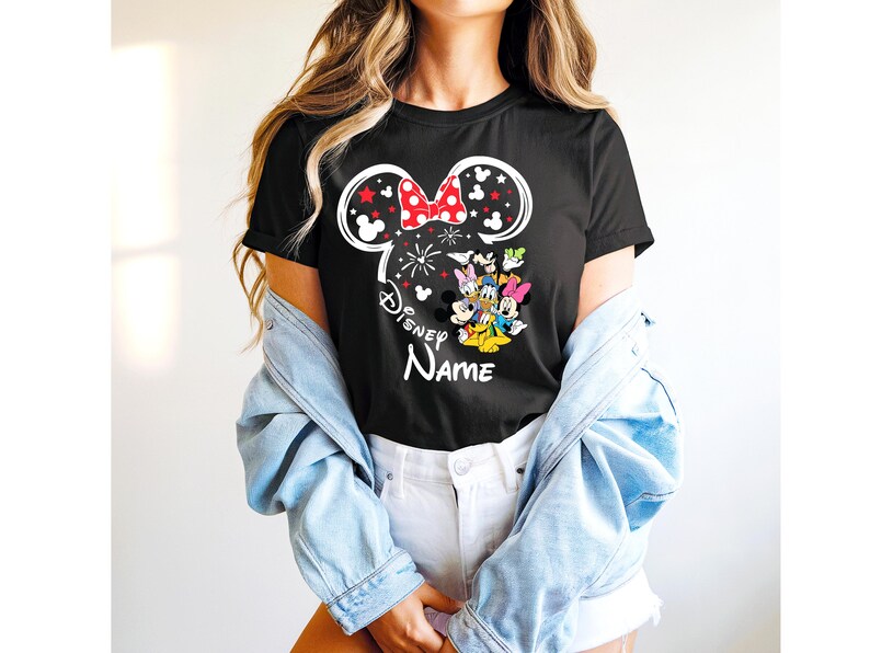 Puede incluir: Camiseta negra con un dise&ntilde;o de Minnie Mouse, un lazo rojo y el texto "Disney Name". El dise&ntilde;o incluye fuegos artificiales y personajes de Disney. La camiseta tiene cuello redondo.