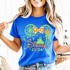 May include: A bright blue t-shirt featuring a neon green and orange graphic of a castle silhouette, a spider web, a pumpkin, and skulls. The design includes a polka-dot bow and the text Disney 2026 with small Mickey Mouse head icons.