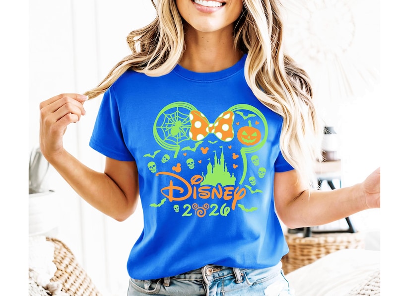 May include: A bright blue t-shirt featuring a neon green and orange graphic of a castle silhouette, a spider web, a pumpkin, and skulls. The design includes a polka-dot bow and the text Disney 2026 with small Mickey Mouse head icons.