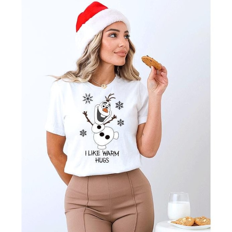 Peut inclure: T-shirt blanc &agrave; manches courtes avec un bonhomme de neige de dessin anim&eacute; et des flocons de neige. Le texte "I LIKE WARM HUGS" est imprim&eacute; en dessous. La personne porte un bonnet de P&egrave;re No&euml;l.