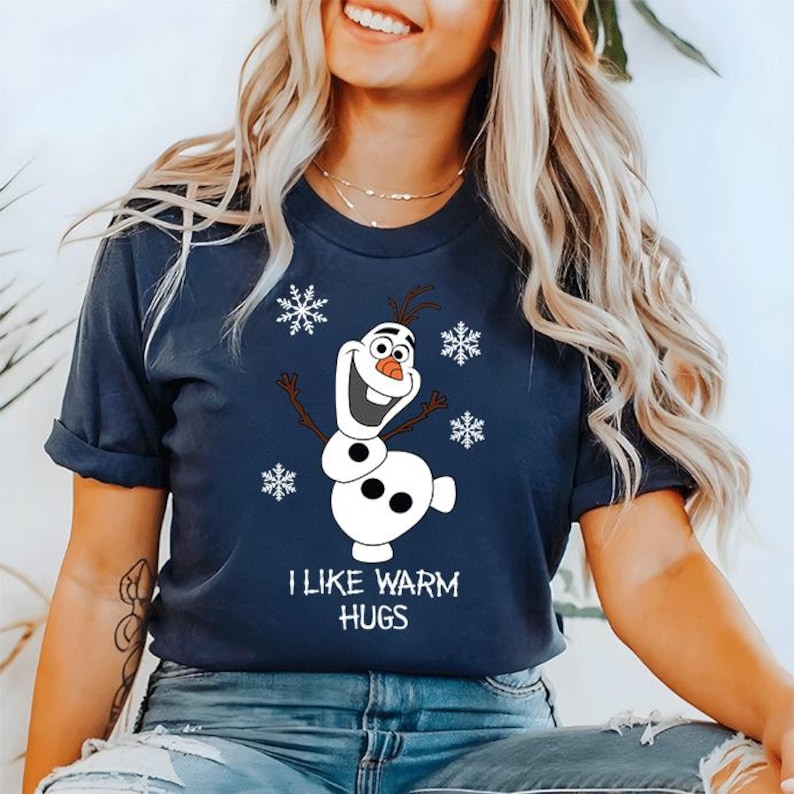 Peut inclure: T-shirt bleu marine avec un bonhomme de neige de dessin anim&eacute; et le texte "I LIKE WARM HUGS" et des flocons de neige. Le bonhomme de neige a un corps blanc, des boutons noirs et des bras en brindilles marron. V&ecirc;tement d&eacute;contract&eacute;.