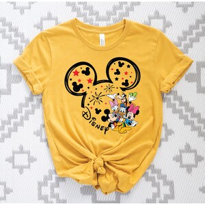 Puede incluir: Camiseta amarilla mostaza con un dise&ntilde;o de la cabeza de Mickey Mouse, fuegos artificiales y estrellas. El dise&ntilde;o incluye a Mickey, Minnie, Donald, Daisy, Goofy y Pluto. La palabra "Disney" est&aacute; escrita debajo.
