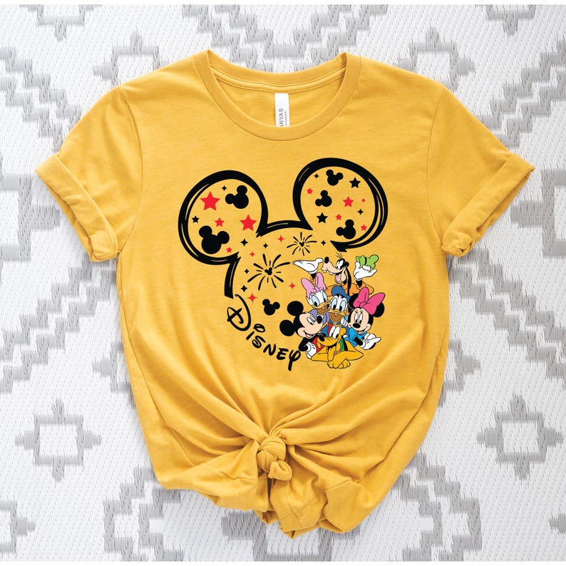 Puede incluir: Camiseta amarilla mostaza con un dise&ntilde;o de la cabeza de Mickey Mouse, fuegos artificiales y estrellas. El dise&ntilde;o incluye a Mickey, Minnie, Donald, Daisy, Goofy y Pluto. La palabra "Disney" est&aacute; escrita debajo.
