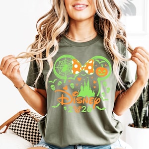 May include: An olive green t-shirt featuring a graphic of a lime green Mickey Mouse ear silhouette, a castle, a spiderweb, a jack-o'-lantern, and small skulls. The design includes an orange polka-dot bow and the orange text Disney 2020.