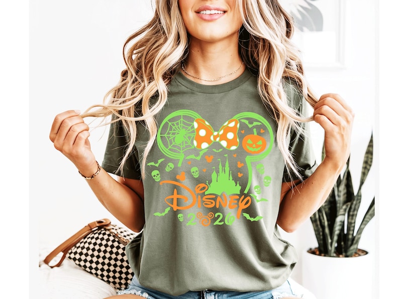 May include: An olive green t-shirt featuring a graphic of a lime green Mickey Mouse ear silhouette, a castle, a spiderweb, a jack-o'-lantern, and small skulls. The design includes an orange polka-dot bow and the orange text Disney 2020.