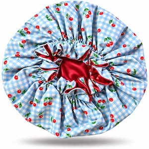 May include: A circular hair bonnet featuring a light blue and white gingham pattern with a repeating print of red cherries and green leaves. The interior of the bonnet is lined with a smooth, solid red fabric, and the edges are gathered with elastic.
