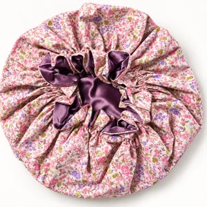 Puede incluir: Un gorro de pelo redondo con un estampado floral de flores rosas, moradas y verdes sobre un fondo blanco. El interior está forrado con una tela lisa de color morado oscuro y los bordes están fruncidos con una banda elástica.