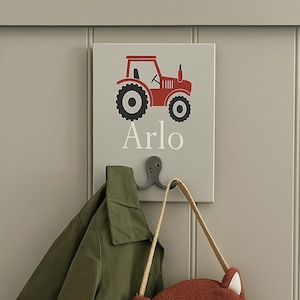 Puede incluir: Un letrero gris montado en la pared con un gráfico de tractor rojo y el nombre "Arlo" en blanco. Debajo, un gancho decorativo sostiene una chaqueta verde y un bolso bandolera con forma de zorro y correa beige.